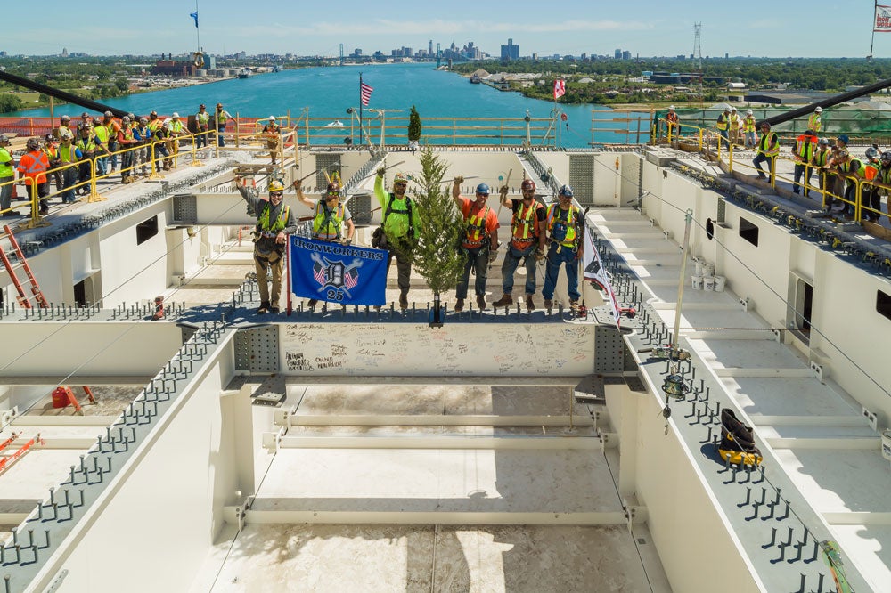 A group of ironworkers stand on the final steel beam, which is signed and decorated with flags and a small tree, ready to be placed on the Gordie Howe International Bridge. The Detroit River and skyline provide a scenic backdrop.