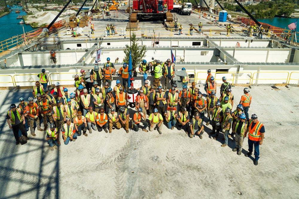 A large group of construction workers in safety gear stand together on the deck of the Gordie Howe International Bridge, marking a milestone in the bridge's construction. The Detroit River and industrial areas can be seen in the background.