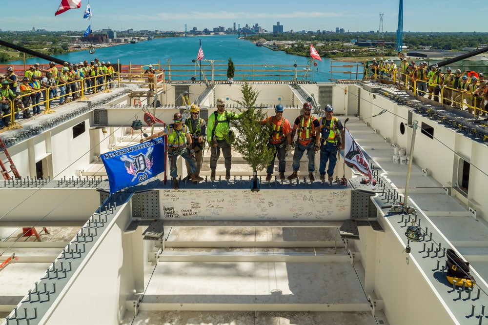 Ironworkers from Local 25 celebrate on the Gordie Howe International Bridge during a topping-out ceremony. The image captures the workers proudly holding their flag on a steel beam, surrounded by signatures, with the Detroit River and city skyline in the background. Safety gear and harnesses highlight the meticulous attention to safety protocols during this milestone in the bridge's construction.